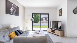 Ein modernes Wohnzimmer mit grauem Sofa, einem Couchtisch mit einem Buch, einem Fernseher auf einem Holzboden und einem großen Fenster mit Blick auf eine grüne Wiese und Gebäude.