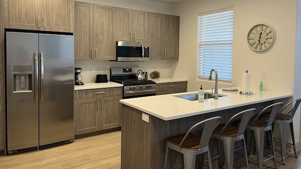 Una cocina moderna con armarios de madera, electrodomésticos de acero inoxidable, una isla central con taburetes y un reloj de pared.