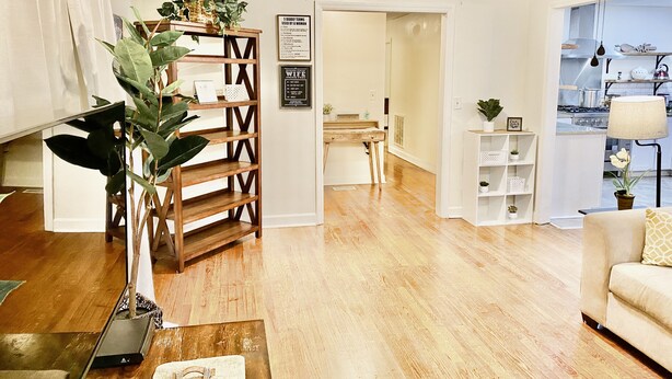 Een moderne woonkamer met houten vloer, een bank, een salontafel en een boekenkast. Op de muur staat een bordje met de tekst 'Welcome' en er is een keuken in de achtergrond zichtbaar.