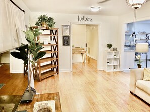 Un salon moderne avec un plancher en bois, un canapé, une table basse et une bibliothèque. Un panneau « Bienvenue » est accroché au mur, ainsi qu’un autre panneau indiquant « Cinq expressions à ne jamais utiliser avec une femme ».