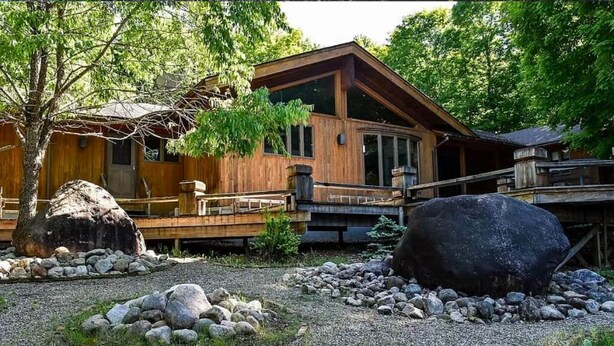 A wooden cabin with a deck, surrounded by rocks and trees. - Luxury Cabin with Indoor Heated SaltWater Pool! (Old Forge)
