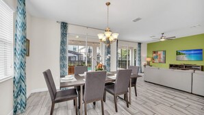 Un espace de salle à manger moderne, avec une table dressée pour quatre personnes, un lustre et une vue sur l’extérieur grâce à de grandes fenêtres.