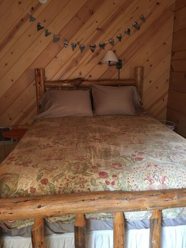 A bedroom with a wooden ceiling, a bed with a floral bedspread, two pillows, and a wall-mounted lamp. - Beautiful Apple Valley Cabin in Sonora on 3.5 Acres walk to Indigeny Ciderworks (Sonora)