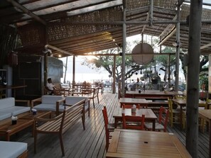 Un restaurante al aire libre con mesas y sillas de madera, un área de estar cubierta y vistas al mar.