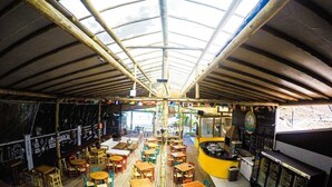 Un restaurant avec un plafond haut, des tables et des chaises en bois, un coin bar et une vue sur l’extérieur.