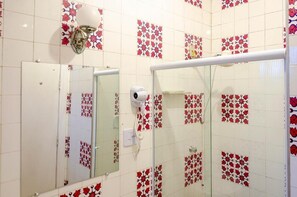 Une salle de bain avec un mur recouvert de carreaux blancs ornés de motifs floraux rouges, une porte blanche, une cabine de douche en verre et un sèche-cheveux fixé au mur.
