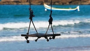 Une scène de plage avec une balançoire suspendue au-dessus de l’eau, un bateau en arrière-plan et des rochers couverts de végétation.