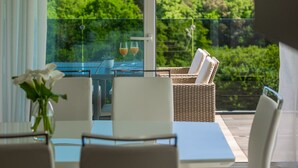 A balcony with a glass table, white chairs, and a wicker chair with cushions. There are two glasses on the table, and a potted plant is visible.