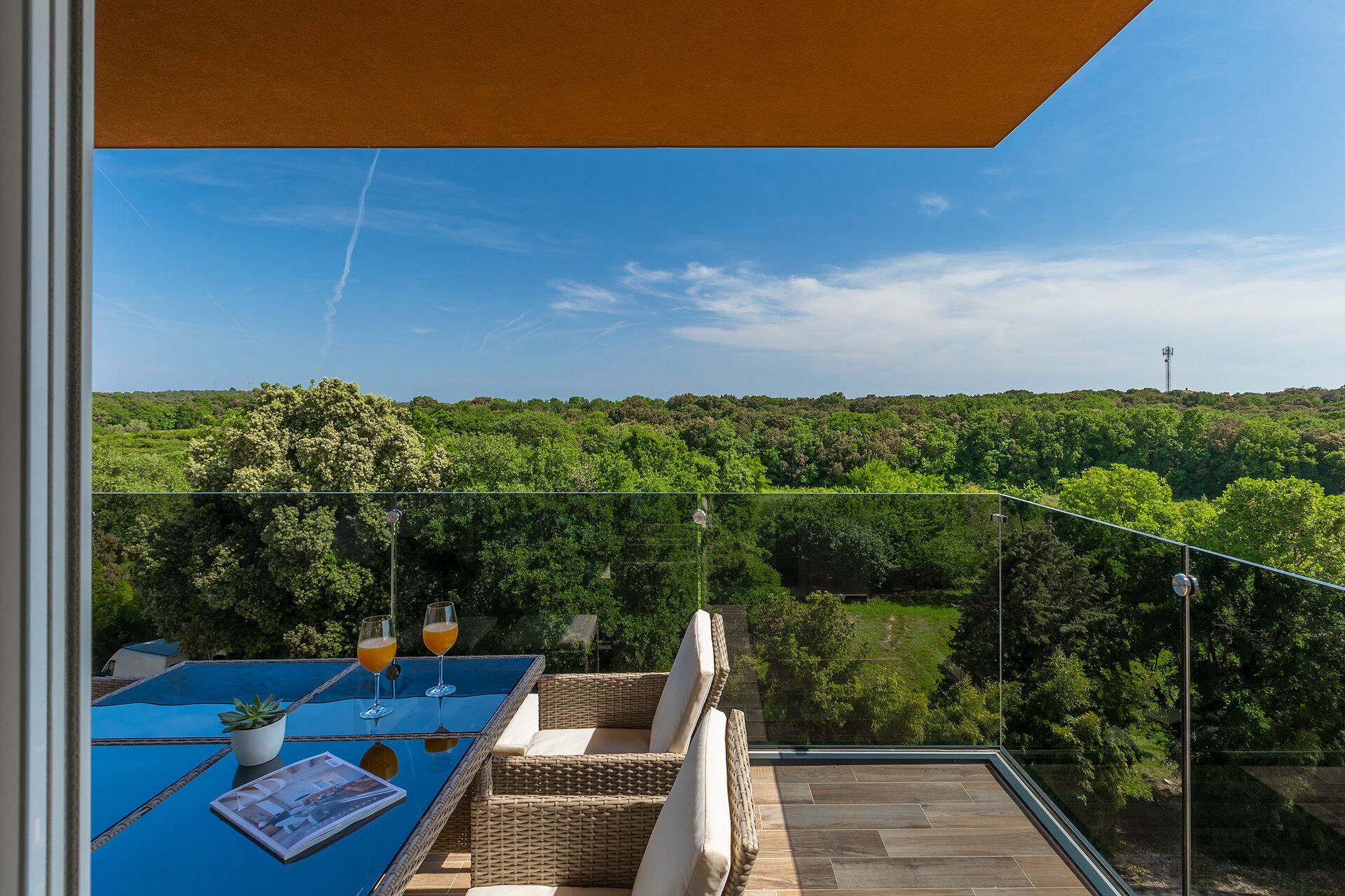 Ein Balkon mit Glastisch, zwei Orangensaftgläsern, einem Blumentopf und Korbmöbeln.