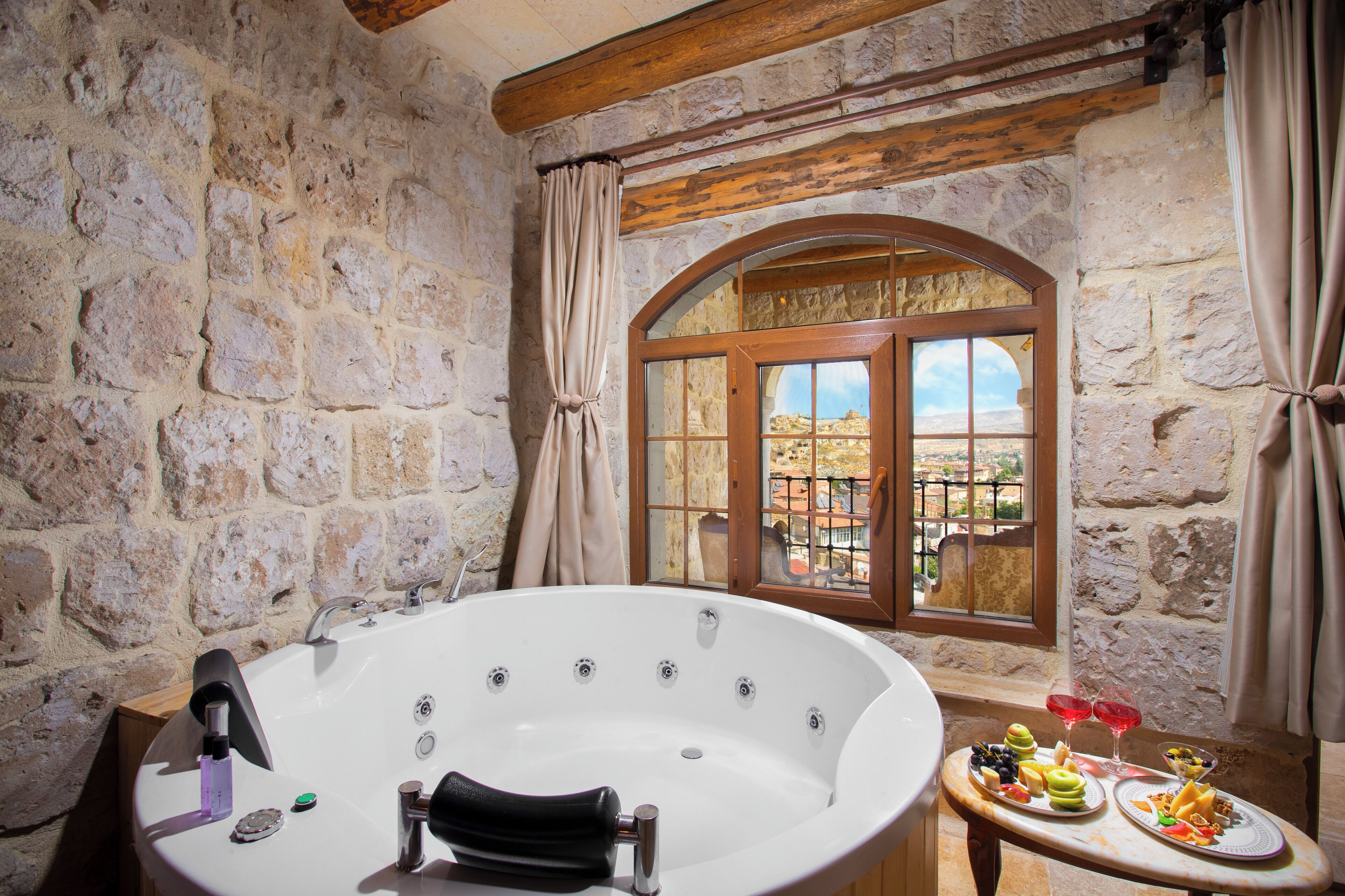 Baño con paredes de piedra, una bañera independiente, una ventana con vistas y una mesa con comida y bebidas.