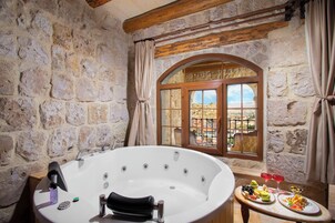 Baño con paredes de piedra, una bañera independiente, una ventana con vistas y una mesa con comida y bebidas.