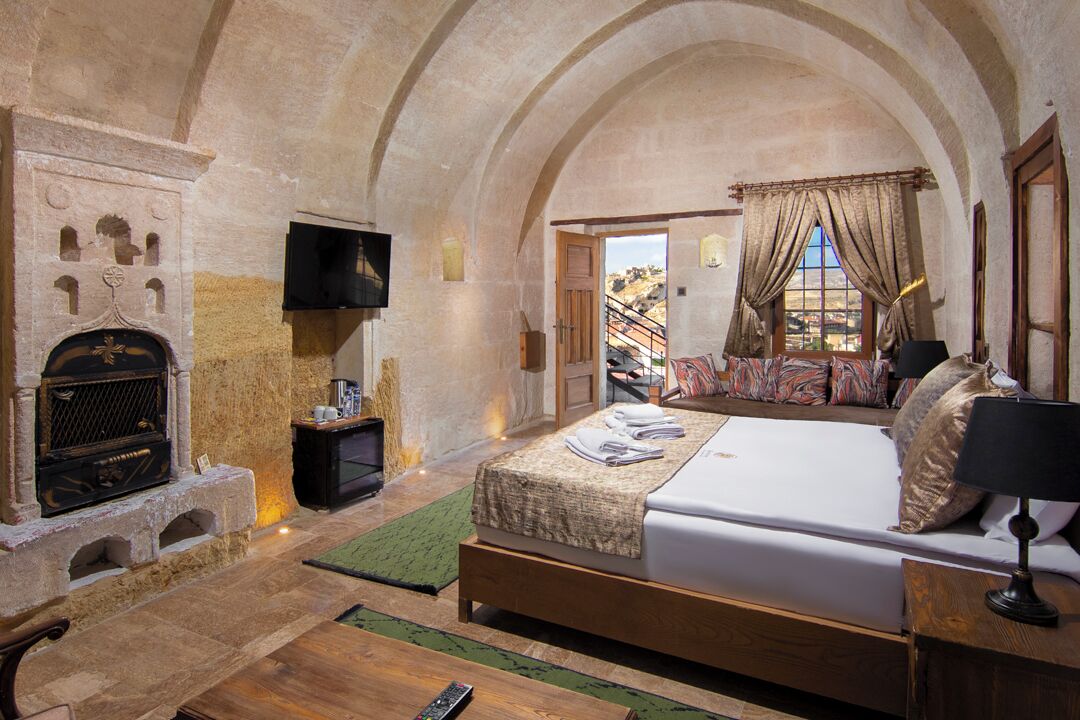 A bedroom with a stone fireplace, a bed with a green rug, and a view of the outside.