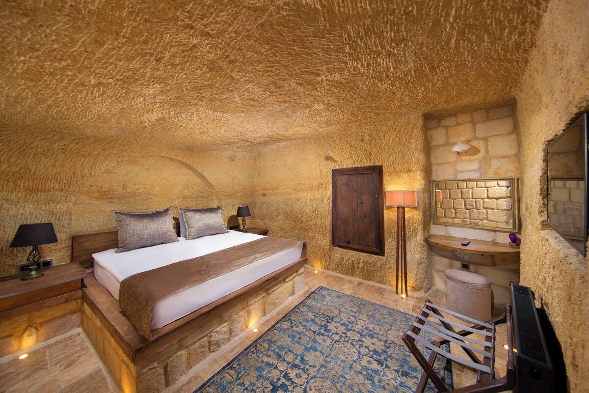 Un dormitorio acogedor y rústico con techo de piedra, cama de madera, mesita de noche con lámpara y un baño pequeño con espejo, además de un tapete azul con estampado.