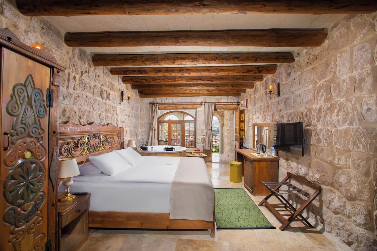 Un dormitorio con paredes de piedra, una cama de madera, una alfombra verde, una mesita de noche de madera, una silla de madera, un aparador de madera, un televisor de pantalla plana y una ventana con cortinas.