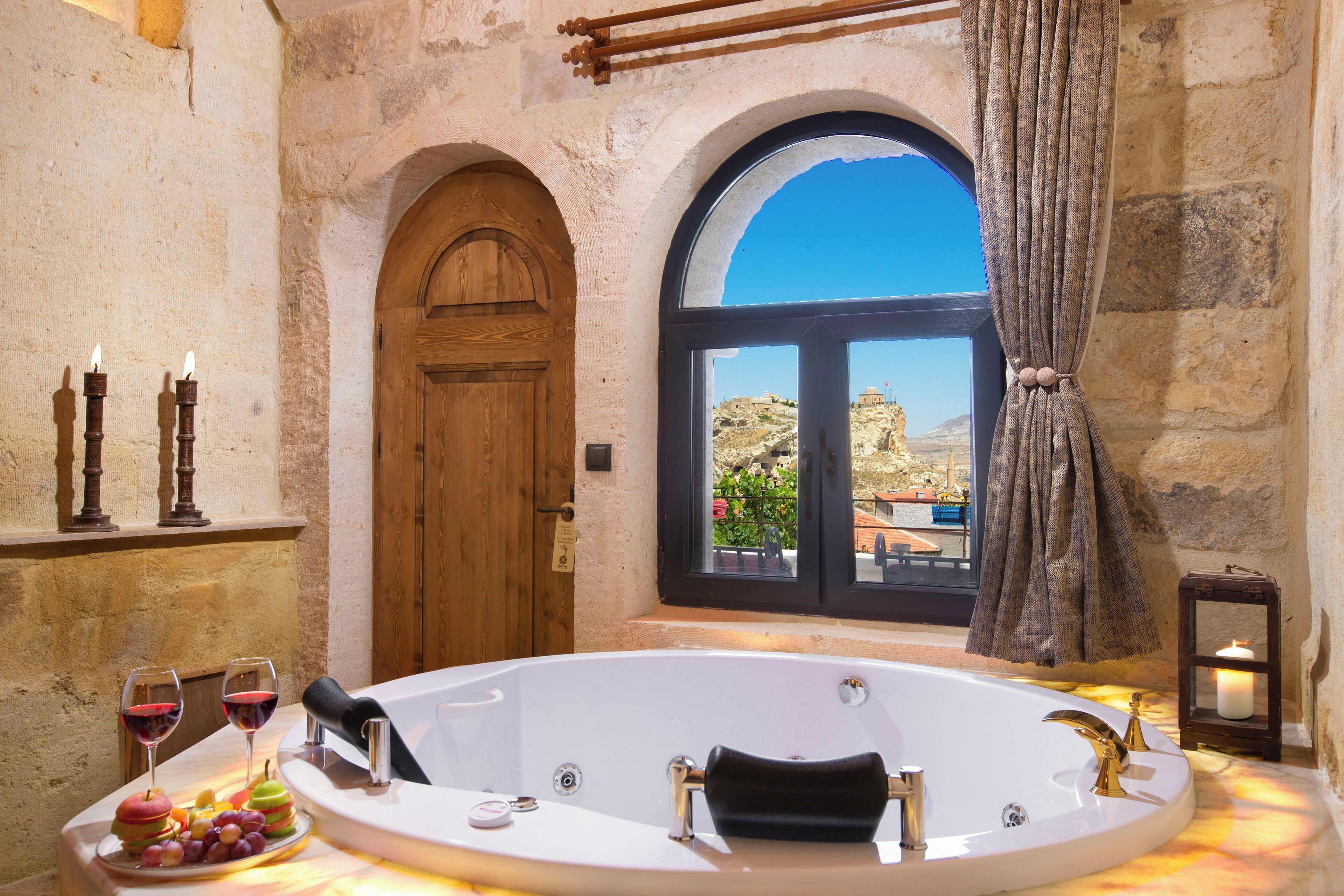 A bathroom with a bathtub, two candles, a window with a view, and a wooden door.