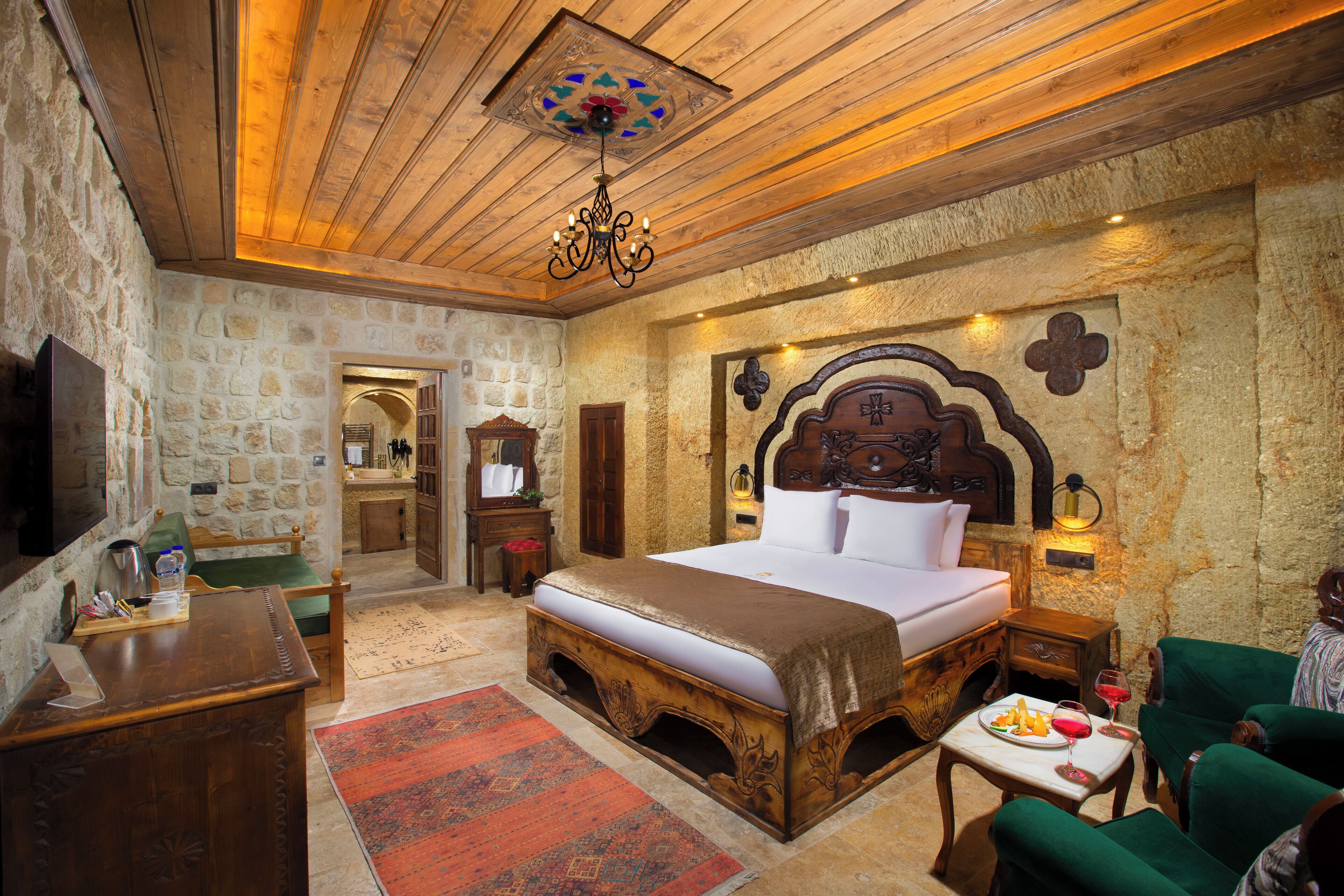 Un dormitorio con pared de piedra, techo de madera, una cama grande, un escritorio de madera, un sillón verde y una pequeña mesa con un plato de comida y una copa de vino.
