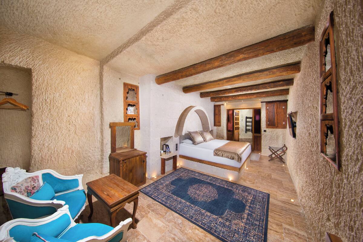 Una habitación con una cama, una mesa de madera, dos sillones azules, un pequeño aparador de madera y una alfombra azul estampada.