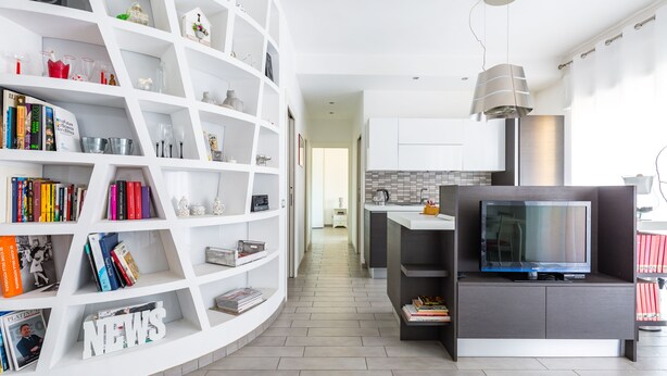 A modern living room with a curved white bookshelf, a flat-screen TV on a dark console, and a kitchen in the background. - MIA - Modern Flat Close Airport and Center (Napoli)
