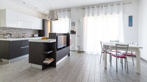 A modern kitchen with a central island, a dining area with a white table and red chairs, and a large window with sheer curtains. - MIA - Modern Flat Close Airport and Center (Napoli)
