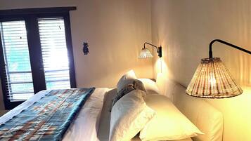 A bedroom with a bed, a bedside lamp, a window with blinds, and a wall-mounted light fixture.