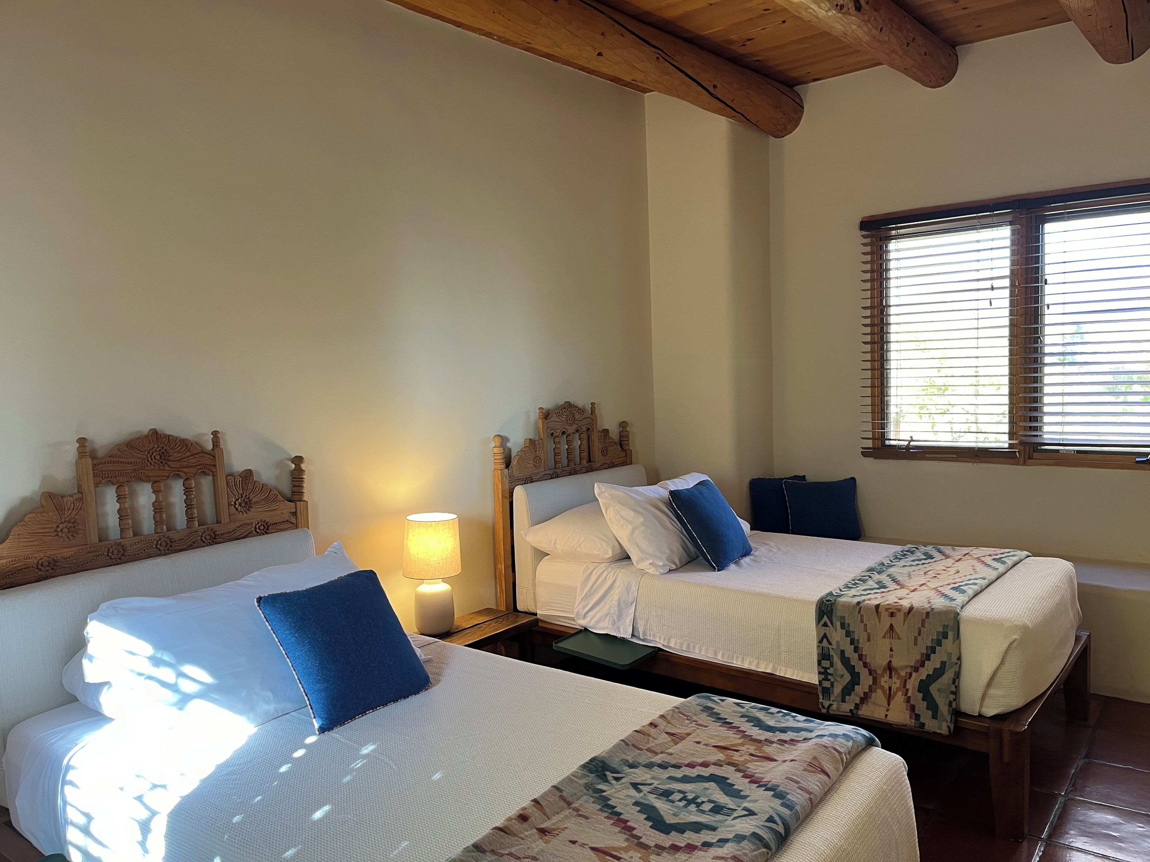 Habitación de hotel con dos camas, cabeceros de madera, una ventana con persianas y una lámpara en una mesita de noche.