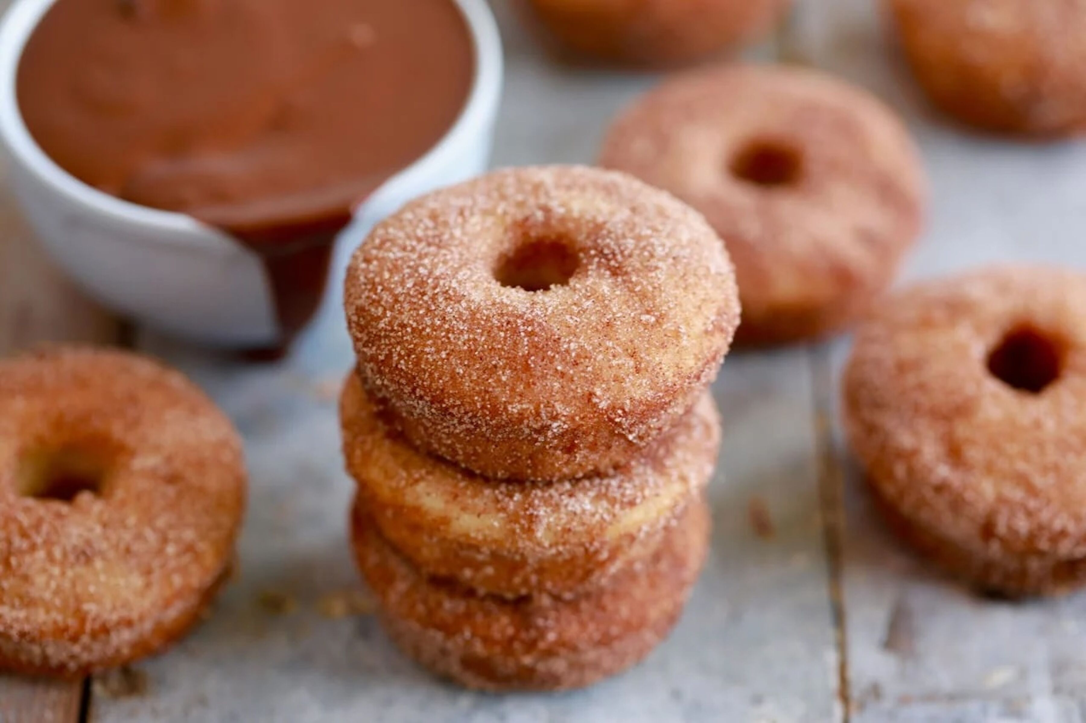 A stack of sugar-coated doughnuts with a chocolate sauce container in the background.