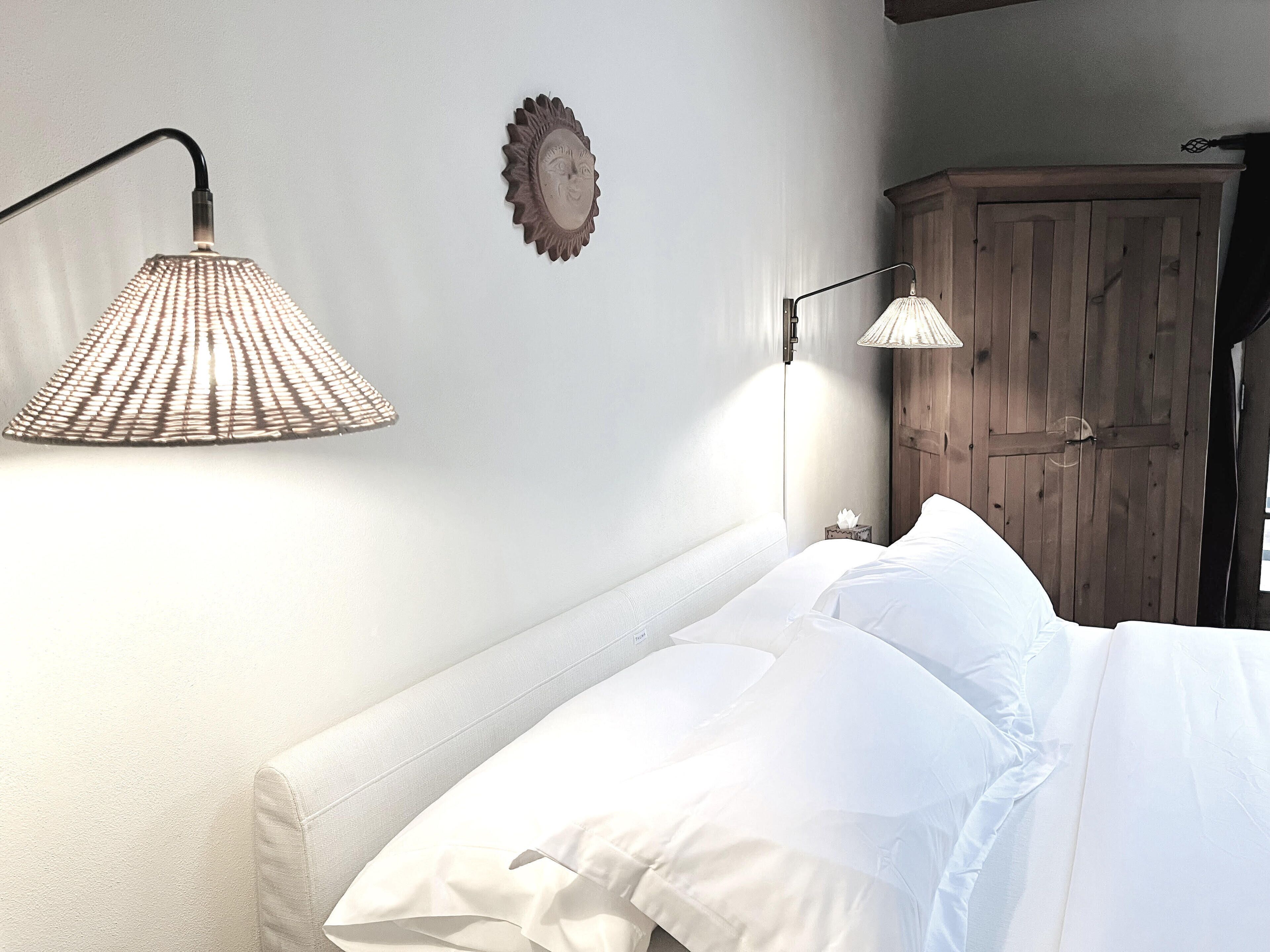 A neatly made bed with white linens, a wooden wardrobe, and two wall-mounted lamps.
