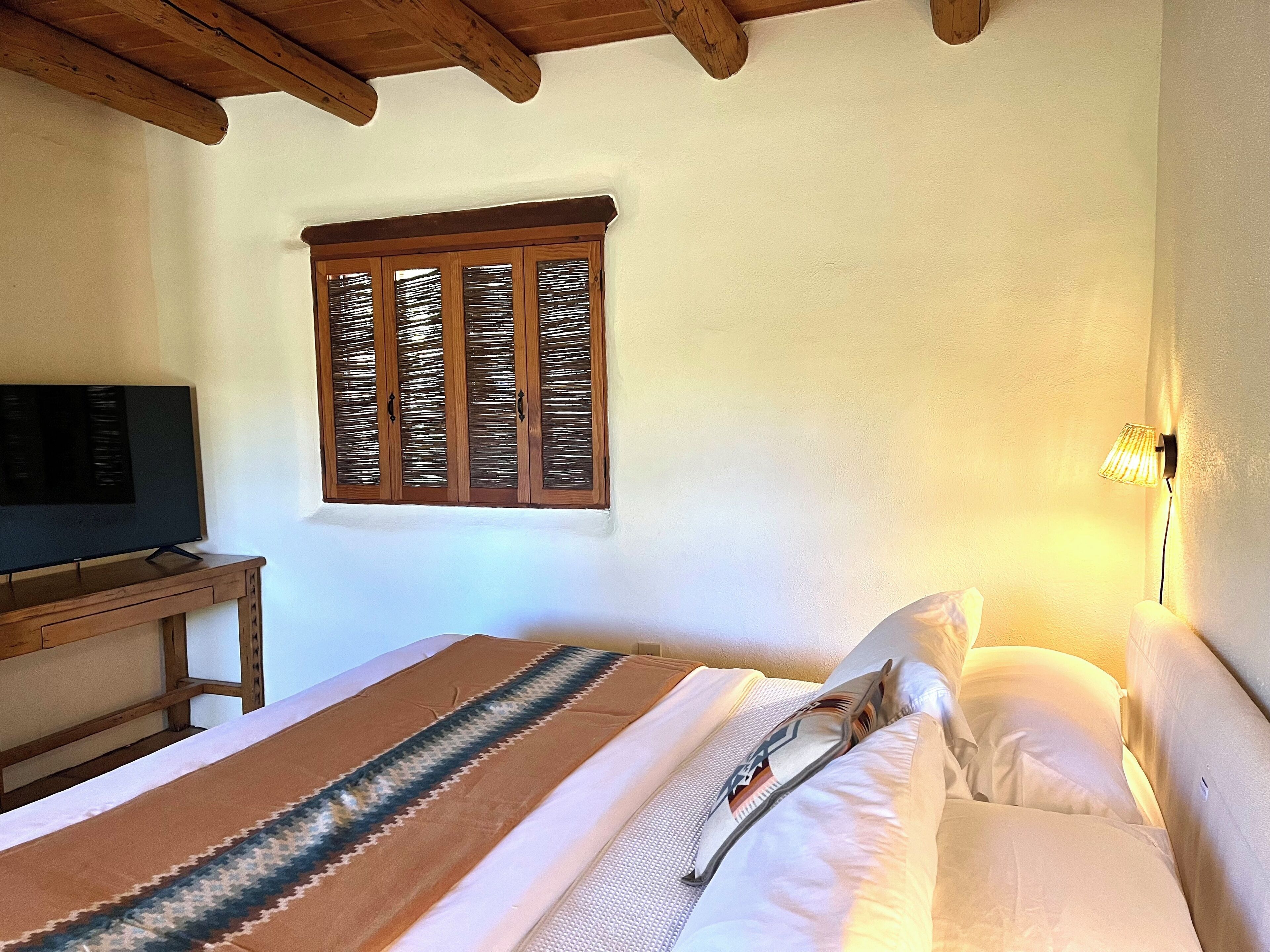 A bedroom with a bed, a television on a wooden table, a window with wooden shutters, and a wall-mounted lamp.