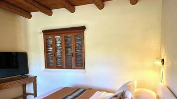 A bedroom with a bed, a television on a wooden table, a window with wooden shutters, and a wall-mounted lamp.