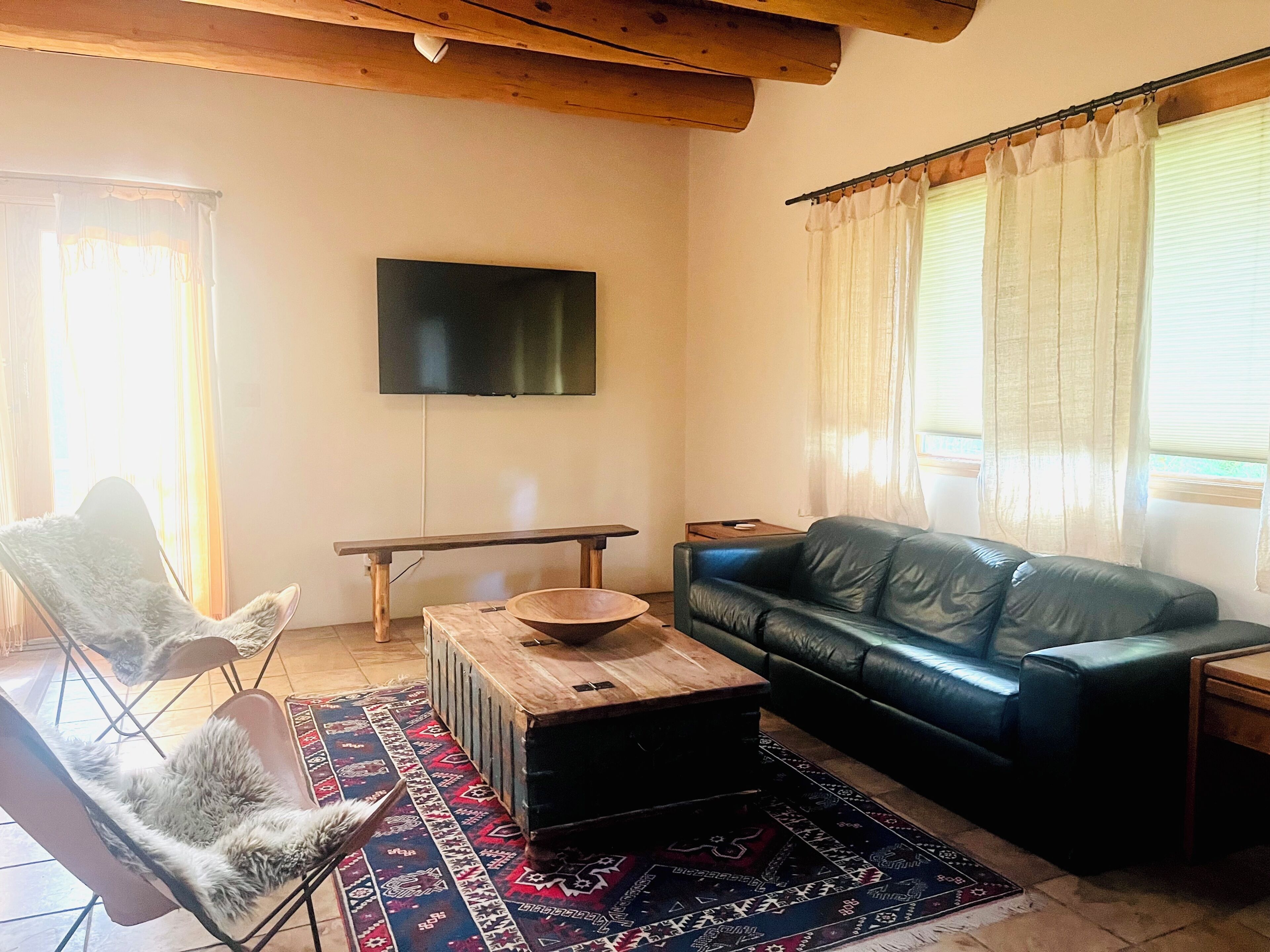 A living room with a black leather sofa, a wooden coffee table, a white furry chair, a flat-screen TV mounted on the wall, and a patterned rug.