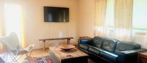 A living room with a black leather sofa, a wooden coffee table, a white furry chair, a flat-screen TV mounted on the wall, and a patterned rug.
