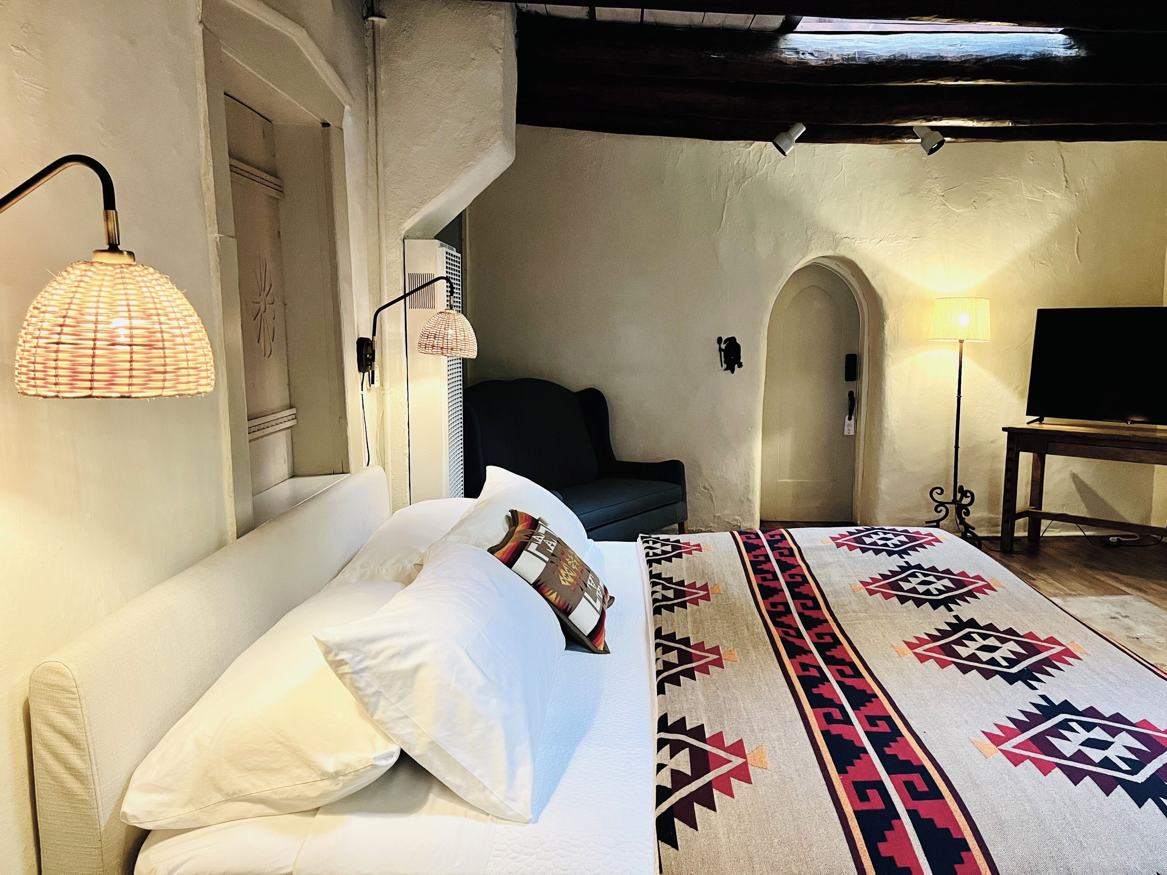 Une chambre à coucher avec un lit, des oreillers, un tapis à motifs, un téléviseur et des lampes fixées au mur.