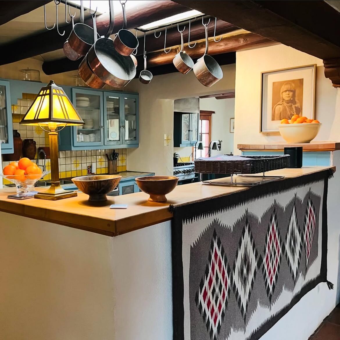A kitchen with hanging pots, a lamp, a bowl of oranges, and a patterned wall.