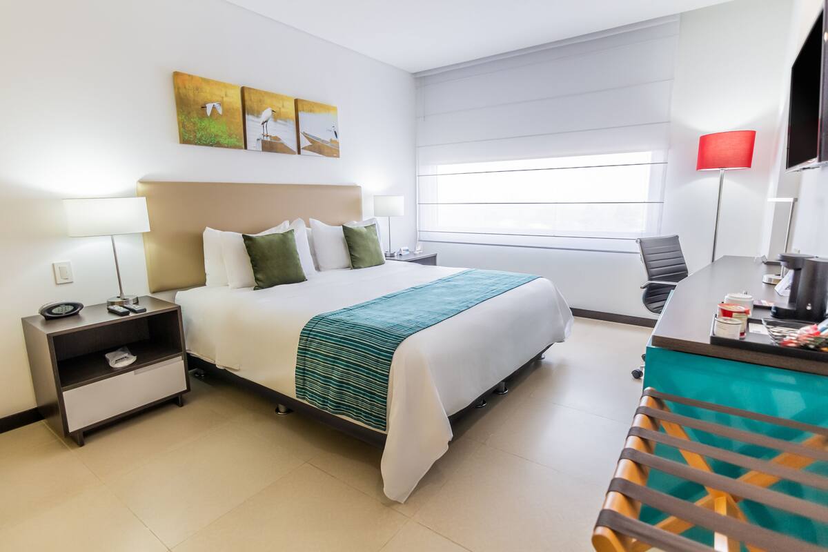 Une chambre d’hôtel comprenant un lit, une table de chevet, une lampe, une œuvre d’art et un bureau avec une chaise.