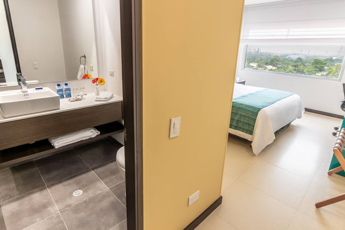Une chambre d’hôtel avec une grande fenêtre, un lit recouvert d’une couverture verte, une salle de bain avec un lavabo et un miroir, et une vue sur la verdure à l’extérieur.