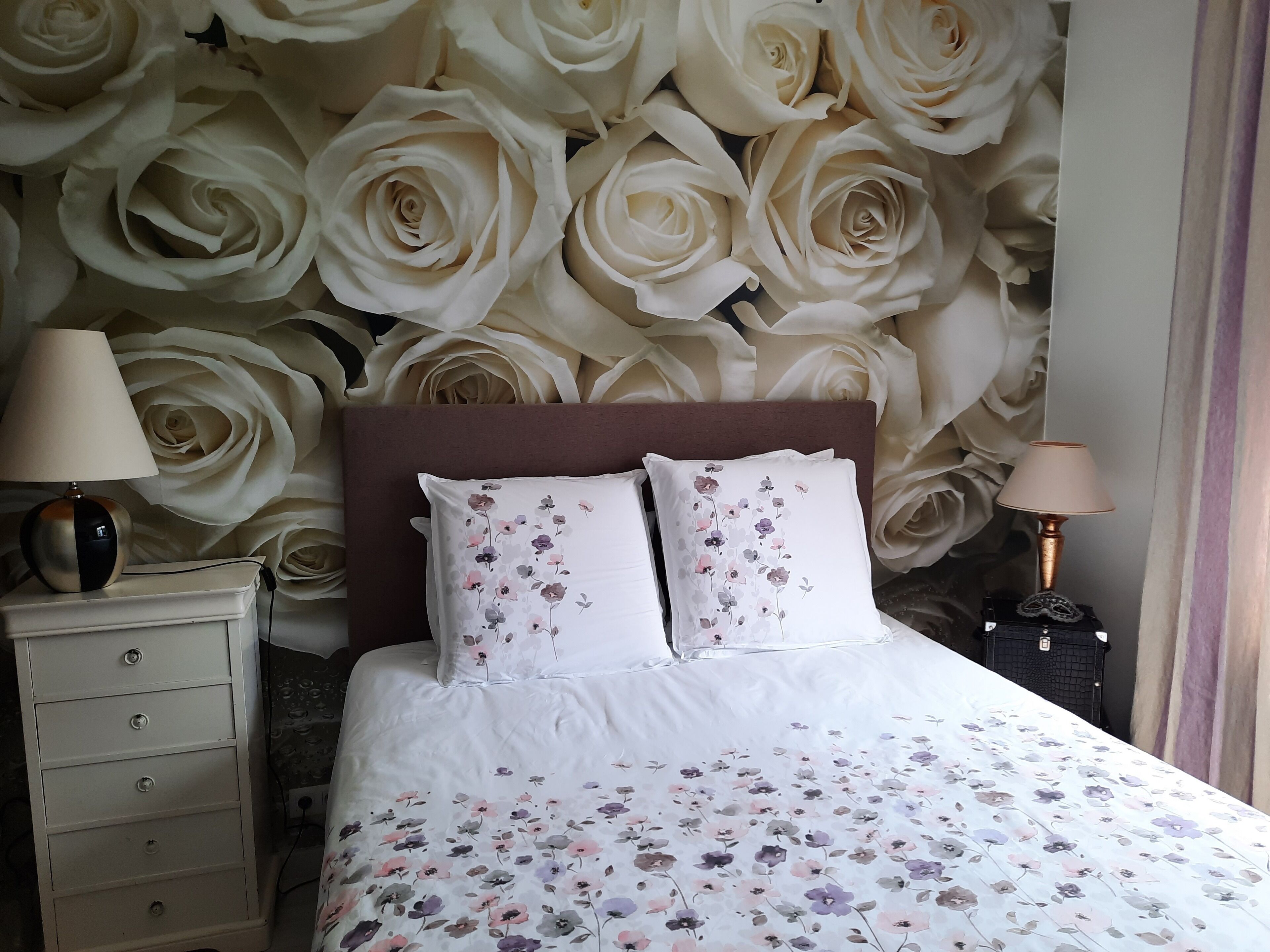 A bedroom with a large floral wallpaper, a bed with floral bedding, two bedside tables with lamps, and a dresser with drawers.