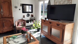 A living room with a television on a wooden cabinet, a chair with a cushion, a glass coffee table, and a large window.