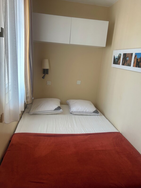 A single bed with two pillows, a red blanket, a wall-mounted lamp, and a framed picture on the wall.