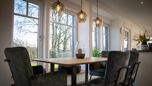 A dining area with a wooden table and grey upholstered chairs, large windows, and hanging pendant lights. - Modern vacation home with terrace and outdoor sauna (Schermbeck)