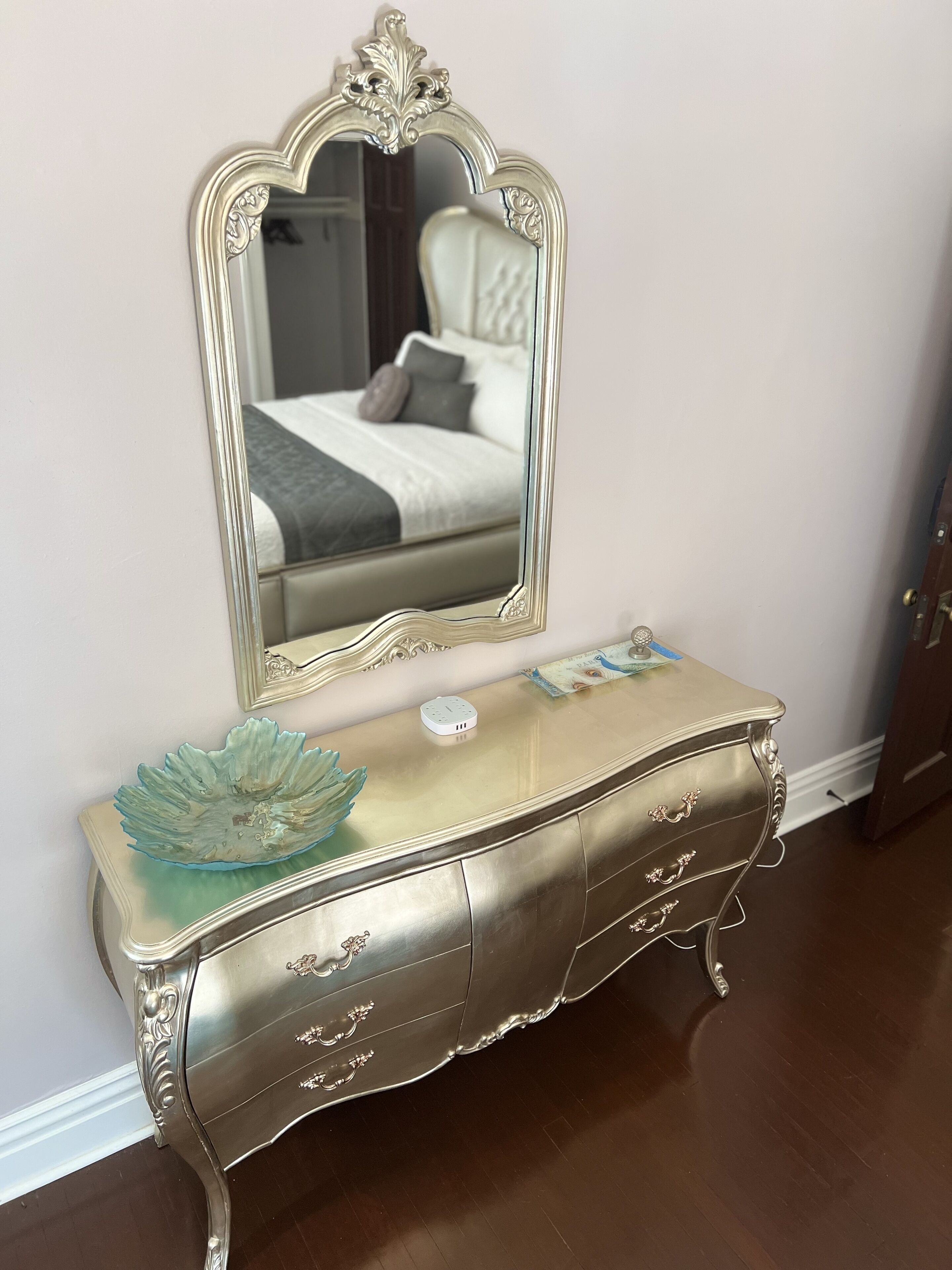 Un aparador de estilo antiguo con tiradores ornamentales y un cuenco decorativo encima, colocado frente a una pared con un espejo grande que refleja un dormitorio.