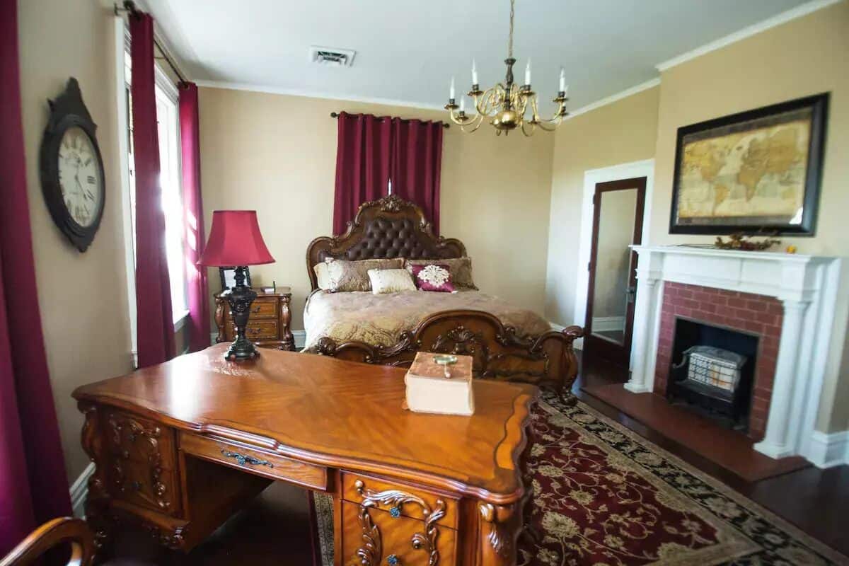 A bedroom with a wooden desk, a bed with a headboard, a chandelier, a fireplace, and a world map on the wall.