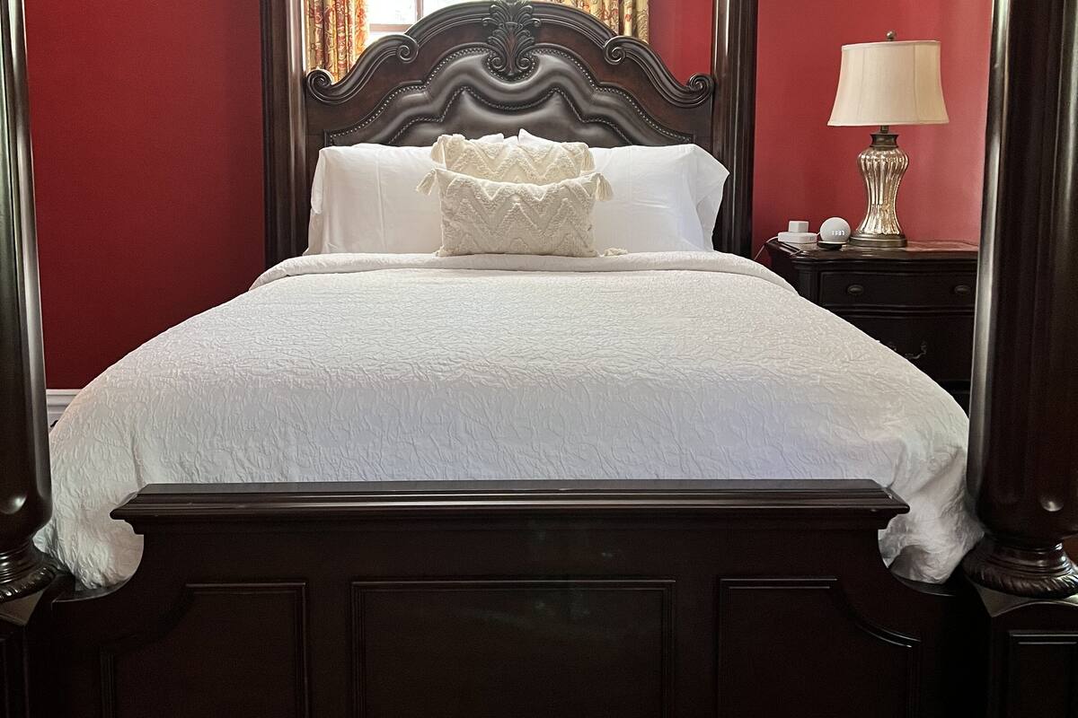 A four-poster bed with a white bedspread and a decorative pillow, set against red walls with a window and curtains in the background.