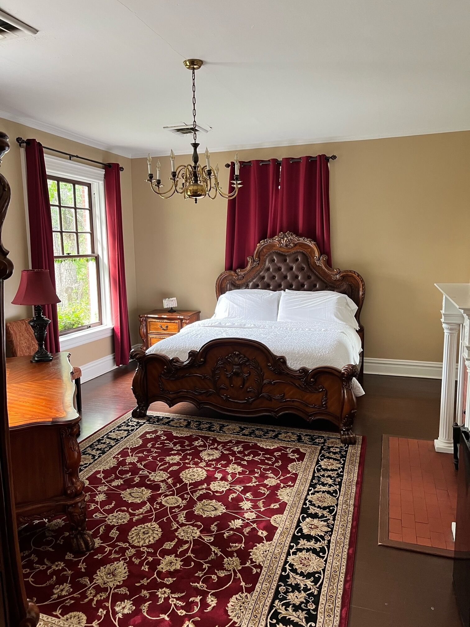 A bedroom with a large bed, a chandelier, a wooden dresser, and a red and black patterned rug.