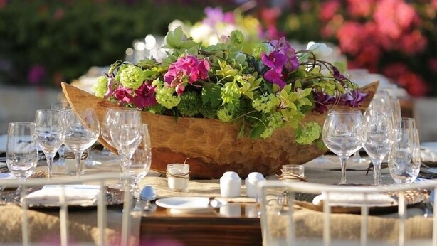 A table set for a meal with a wooden centerpiece filled with flowers, surrounded by empty wine glasses and plates.