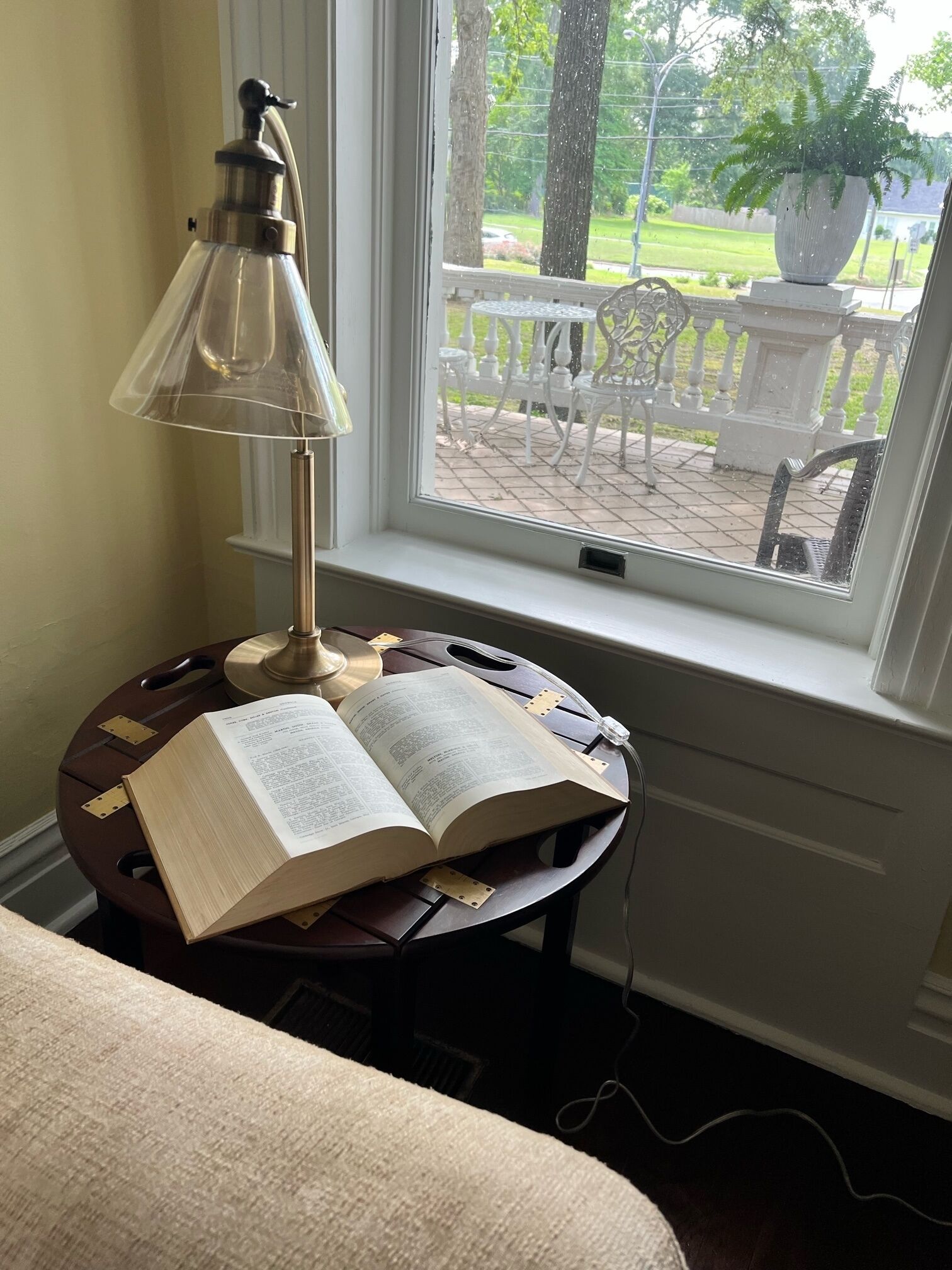 Un tavolo rotondo in legno con una lampada e un libro aperto, posto vicino a una finestra che si affaccia su un recinto bianco e sulla vegetazione.