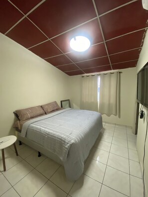 A bedroom with a bed, a small table, a TV, and a ceiling light.