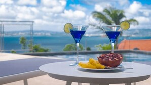 A table with two martini glasses, a bowl of fruit, and a pomegranate on a balcony overlooking a body of water and a cityscape. - Tangarane offers two luxury villas with private pools and exceptional views over (LES TROIS ILETS)