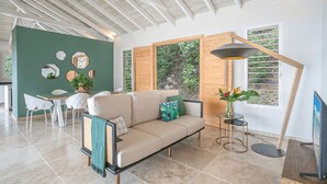 A modern living room with a beige sofa, a dining table with chairs, a floor lamp, and a large window with a view of greenery. - Tangarane offers two luxury villas with private pools and exceptional views over (LES TROIS ILETS)