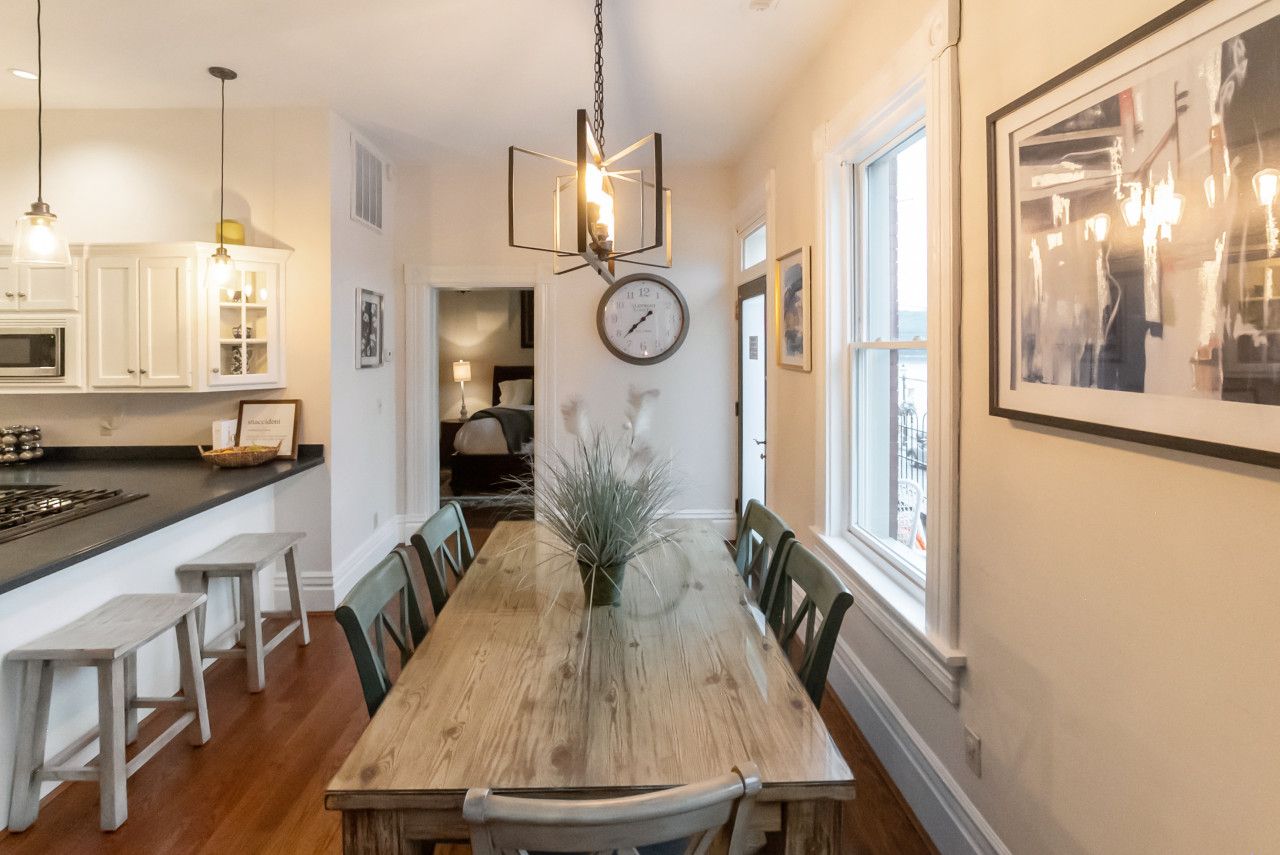 A dining area with a wooden table, chairs, a chandelier, a clock, and a view of the kitchen.