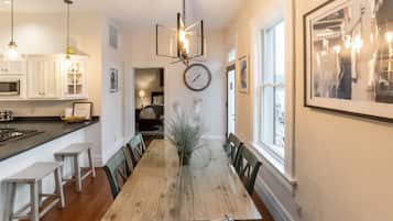 A dining area with a wooden table, chairs, a chandelier, a clock, and a view of the kitchen.